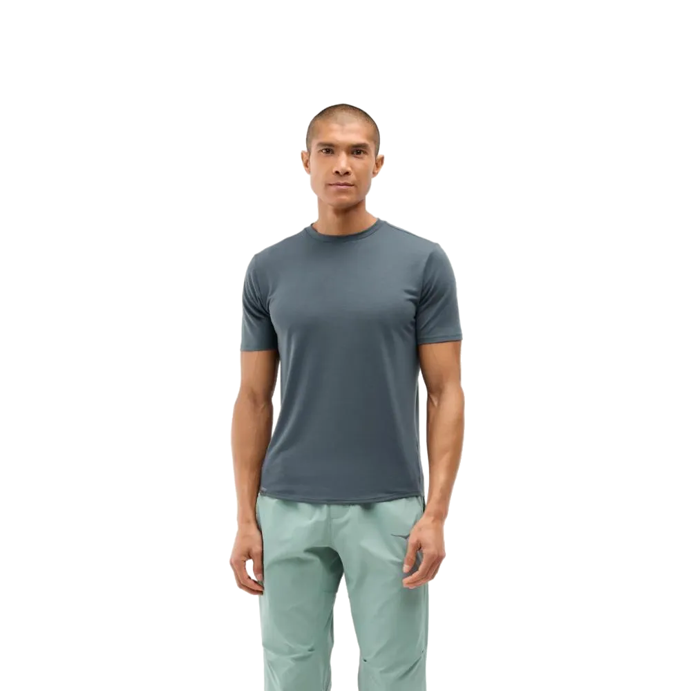גבר עומד עם פניו קדימה, לבוש בחולצת ה-Hoka 1141757/FGYN M HOKA ESSENTIAL TEE בצבע Foggy Night מבית Hoka, בשילוב מכנסיים בצבע ירוק בהיר, על רקע לבן חלק.