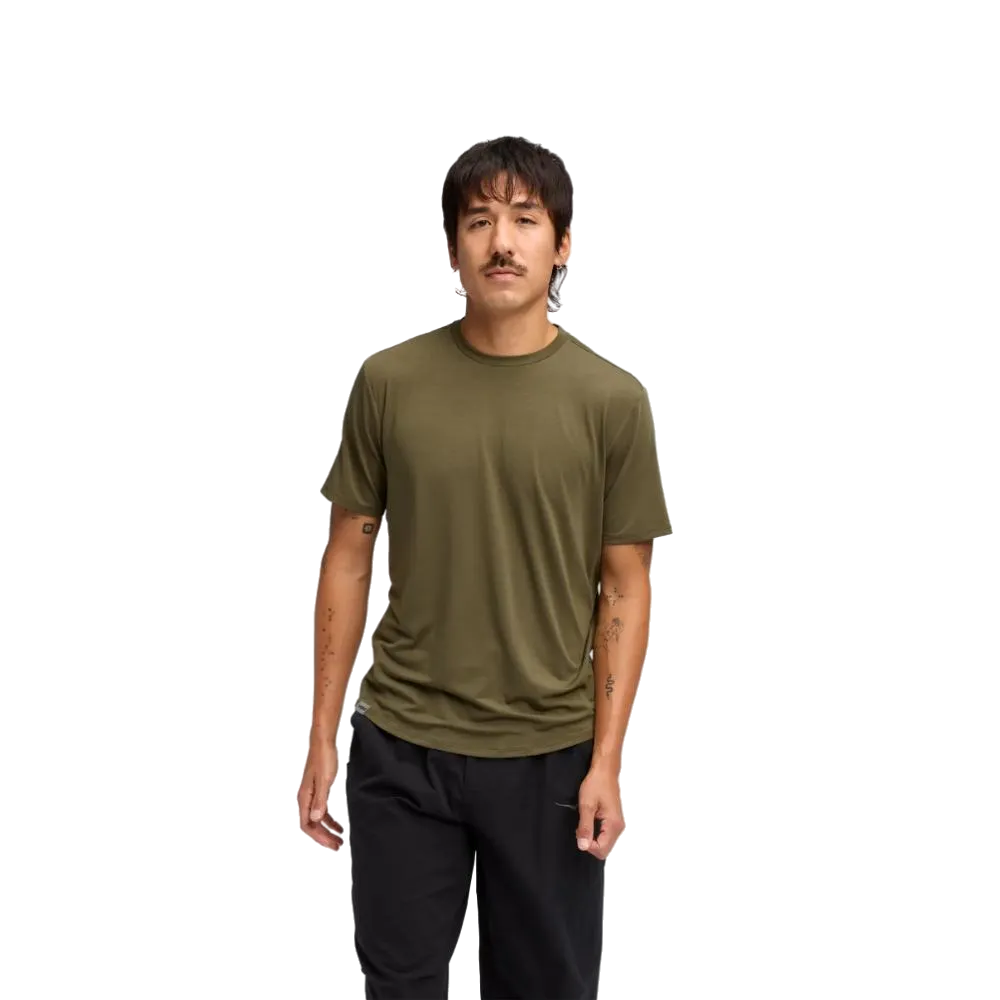 גבר עם שפם, לבוש בחולצת הוקה Essential בצבע זית עתיק (Hoka 1141757/ATQL), עומד בביטחון על רקע לבן חלק.