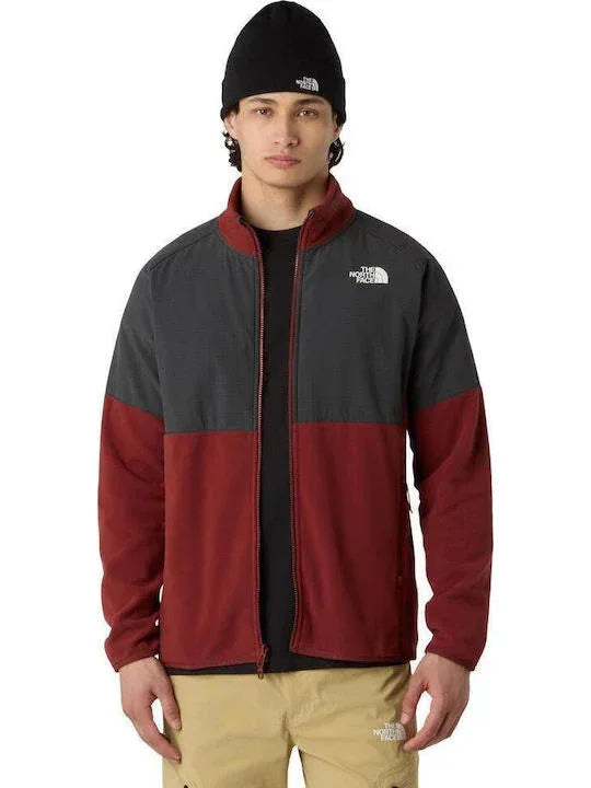 אדם עומד עם הפנים קדימה על רקע אחיד, לבוש בכובע צמר שחור, מכנסיים חומים וז'קט The North Face GLACIER HEAVYWEIGHT FULL ZIP TA89JGDO3 בצבעים אדום ואפור.