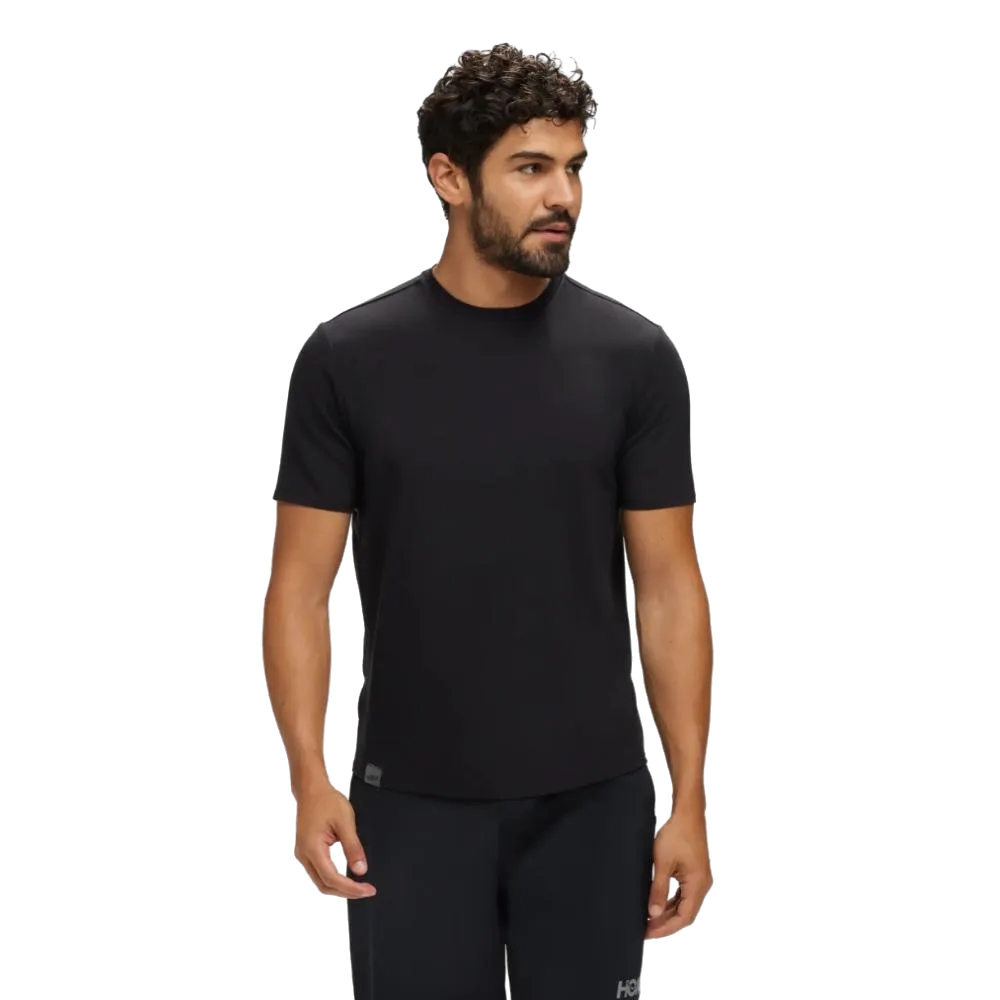 גבר עם שיער קצר ומתולתל לובש את חולצת ה-HOKA ESSENTIAL TEE בדגם 1141757/BLK בצבע שחור מבית Hoka ומכנסיים כהים, כשהוא עומד על רקע לבן פשוט.