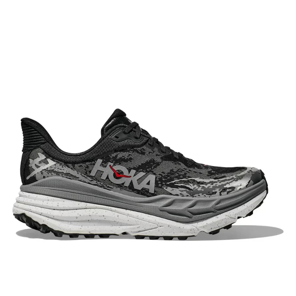 ה-Hoka STINSON 7 BLACK/OUTER ORBIT (1141530/BCKT) הוא נעל ריצה בצבעי שחור ואפור עם חלק עליון בדוגמת הסוואה, סוליה אמצעית מנוקדת בלבן וסוליה חיצונית עמידה בצבע שחור, כפי שמוצג בפרופיל הצדדי.