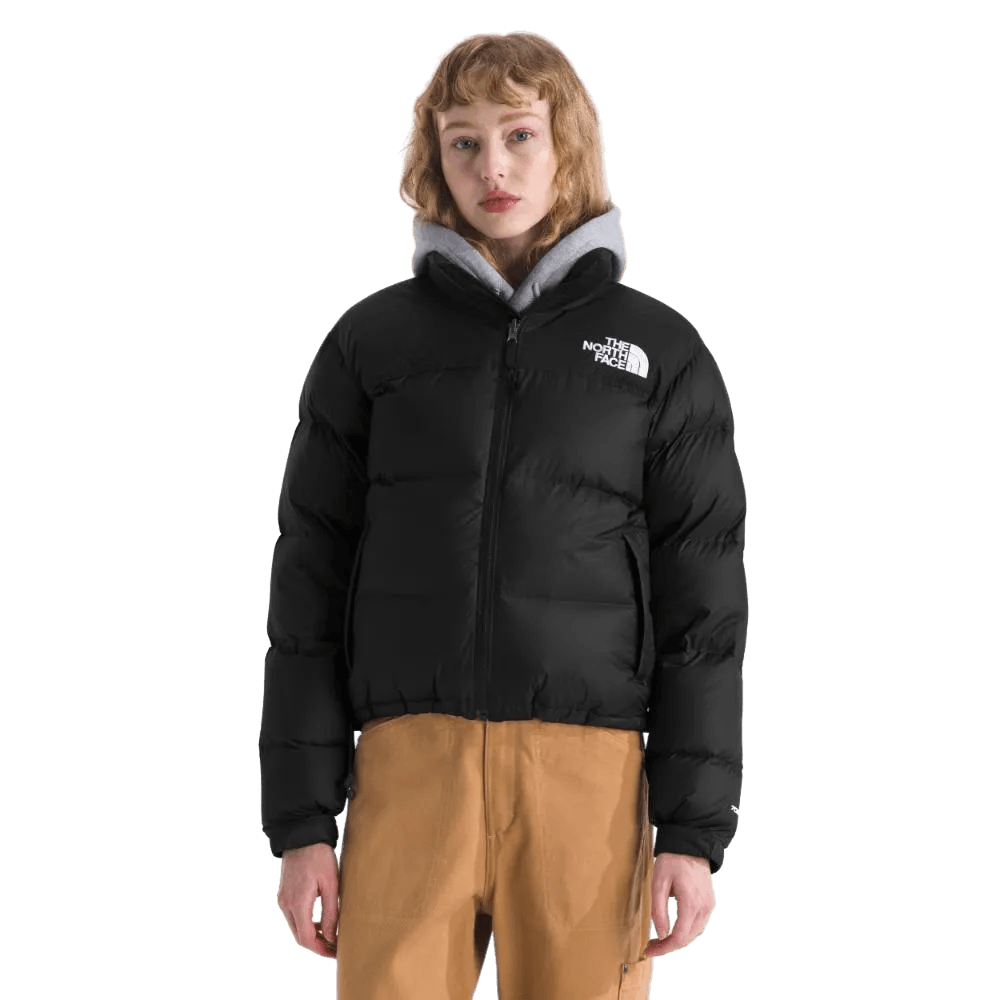 אדם לבוש במעיל שחור The North Face 1996 RETRO NUPTSE JACKET TA3XEOGOF מעל קפוצ'ון אפור ומכנסיים חומים עומד על רקע לבן.
