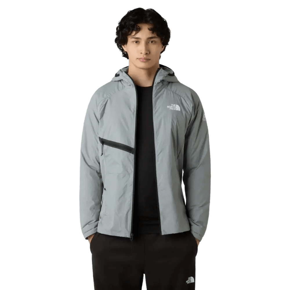 גבר לובש ג'קט אפור בהיר של The North Face עם קפוצ'ון גברים MOUNTAIN ATHLETICS INSULATED HOODED JA TA8EJ7H5F מעל חולצה שחורה ומכנסיים שחורים, עומד על רקע לבן חלק.