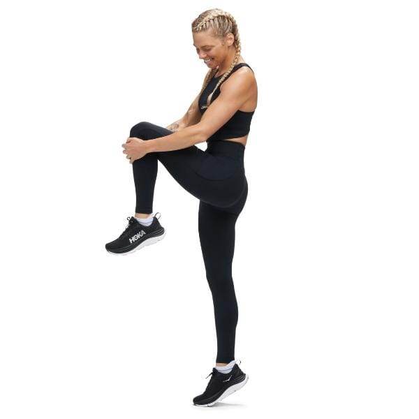 אדם שלובש את Hoka 1141763/BLK W Elaro Knit Tight 27" מבצע תרגיל הרמת ברכיים על רגל אחת על רקע לבן רגיל.