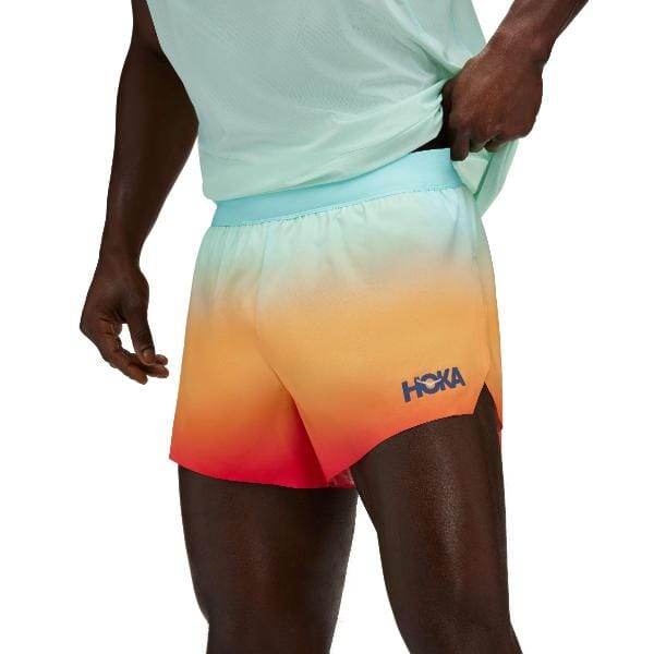 אדם לובש חולצה ירוקה בהירה ו-Hoka 1155134/CDLS Race Day Split Short הכולל את הלוגו "HOKA", התאמת חולצתו.