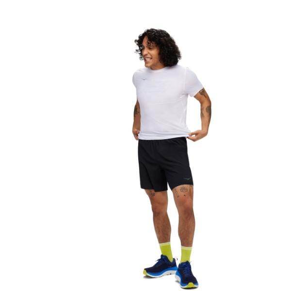 אדם שלובש חולצה לבנה, מכנסי GLIDE 7'' SHORT 2IN1 שחורים של Hoka ונעלי ריצה כחולות עומדים על רקע לבן.