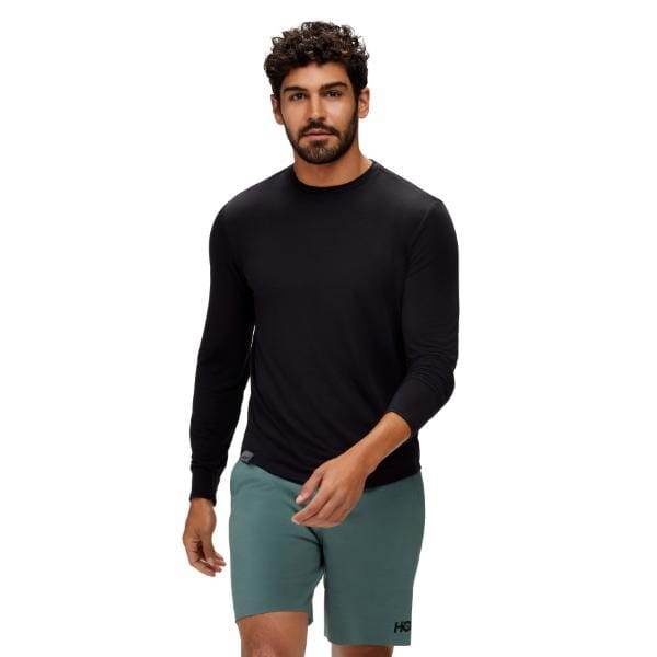 גבר שלובש את Hoka Essential LS Tee בשחור בשילוב עם מכנסיים קצרים ירוקים הולך קדימה על רקע לבן פשוט.