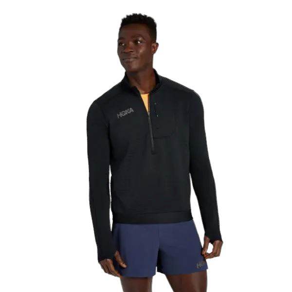 אדם שלובש מכנסיים קצרים של Hoka 1135054/BLK M 1/2 Zip וכחול עומד על רקע לבן.